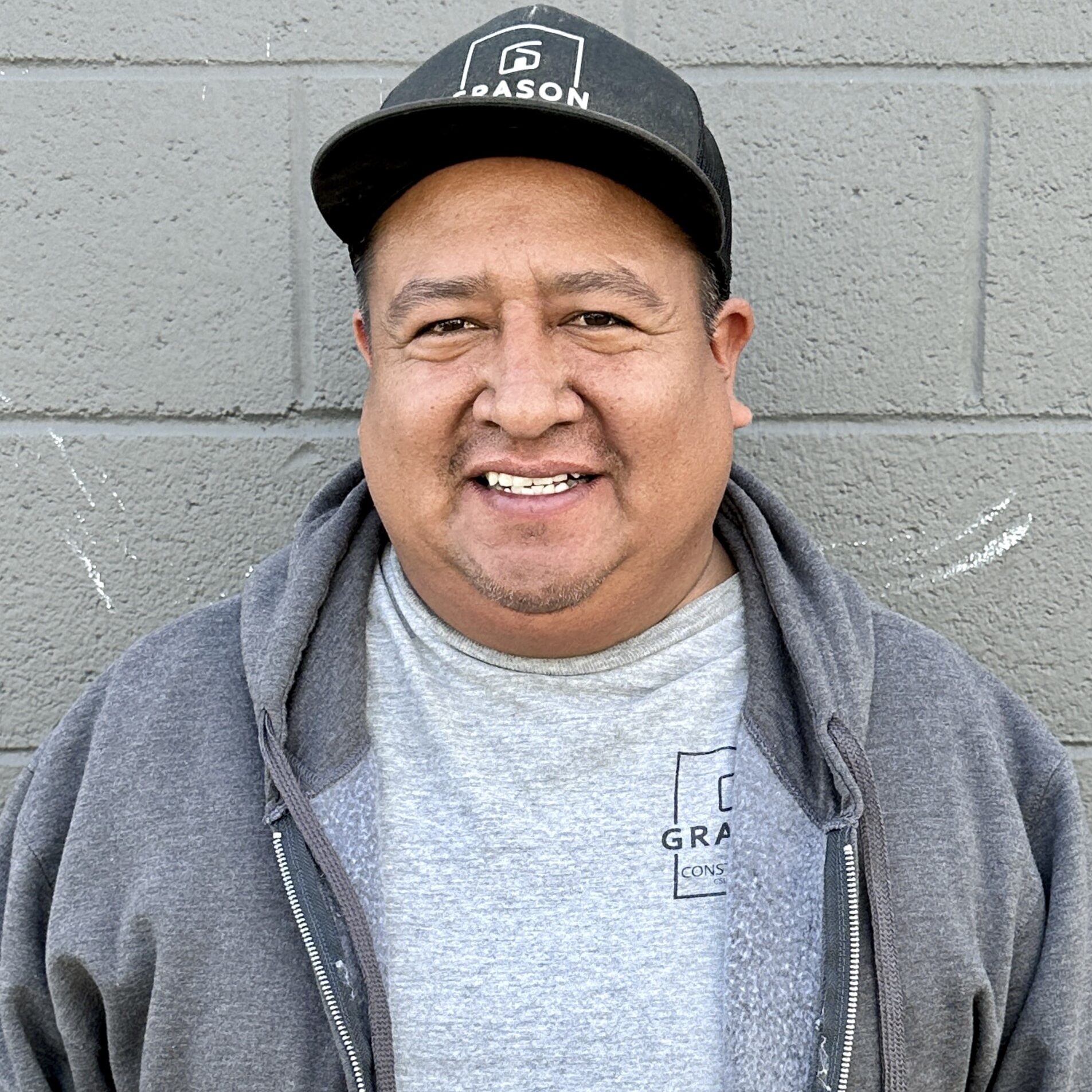 A man wearing a black cap and grey hoodie smiling in front of a grey wall.